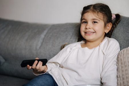 Portrait of cute little girl using remote controller of TV and watching cartoons. Small but already so grown upの写真素材