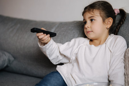Portrait of cute little girl using remote controller of TV and watching cartoons. Small but already...