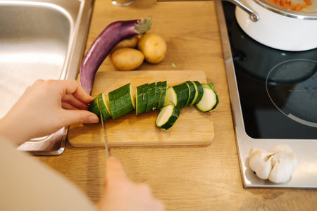 First person view of female hand cutting green zucchini on wooden table at the kitchen. Home food...