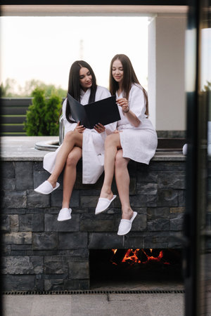 Portrait of two gorgeous woman in white bathrobe sits in front of hot tube outdoors. Sexy lady on luxury resortの写真素材
