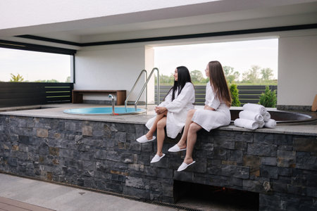 Two beautiful woman in white bathrobe sits in front of hot tube outdoors. Sexy lady on luxury resortの写真素材