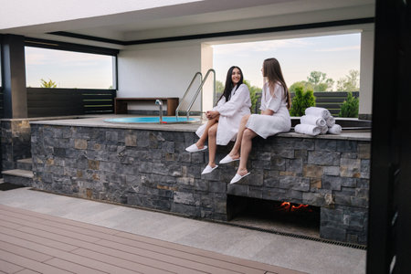 Two beautiful woman in white bathrobe sits in front of hot tube outdoors. Sexy lady on luxury resortの写真素材