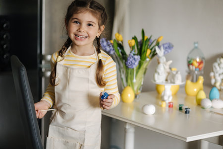 Cute four year girl in beige apron sitting on kitchen table and painting easter eggsの写真素材