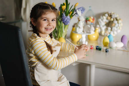Cute four year girl in beige apron sitting on kitchen table and painting easter eggsの写真素材