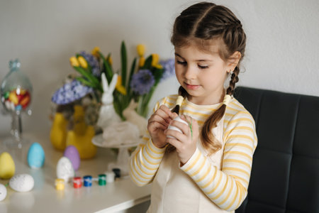 Adorable little girl painting eggs on kitchen at home. Cute girl in apron. Background of easter decorationの写真素材