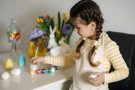 Adorable little girl painting eggs on kitchen at home. Cute girl in apron. Background of easter decorationの写真素材