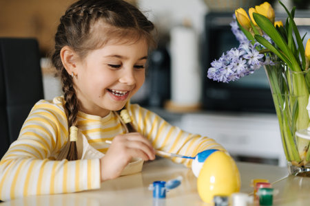 Cute four year girl sitting on kitchen table and painting easter eggs. Yellow and blue colour. Ukrainian symbolicの写真素材