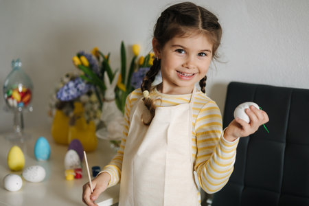 Adorable smiled girl hold brush and eggs in hands and preparing for painting. Easter decoration on kitchenの写真素材