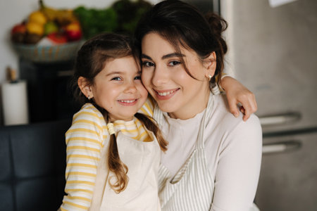 Close-up portrait of daughter with mom on kitchen. Beautiful girls dressed in apronの写真素材