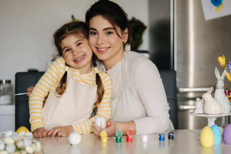 Portrait of adorable little girl with mom on kitchen. Beautiful woman help her daughter paint easter eggsの写真素材