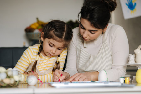 Mom and her cute little girl sitting at the table in the kitchen and color the picture with pencilsの写真素材