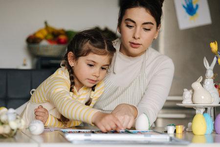 Mom and her cute little girl sitting at the table in the kitchen and color the picture with pencilsの写真素材