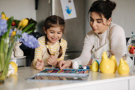 Mom helping her daughter paint an Easter bunny mask. Adorable little girl sitting at the table in the kitchenの写真素材