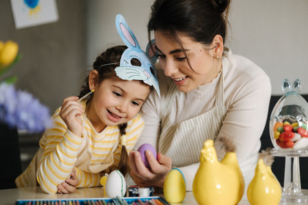 Easter time with family. Adorable girl wore bunny mask and help mom painting eggs. Spring decoration in kitchenの写真素材