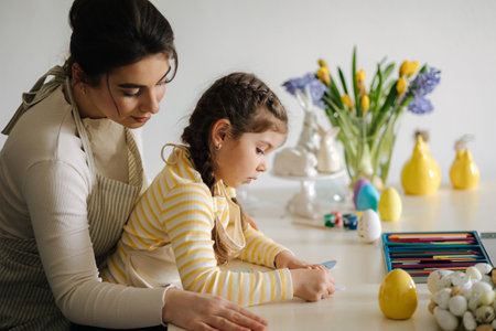 Side view of mom helping her daughter paint an Easter bunny mask. Little girl sitting at the table in the kitchenの写真素材