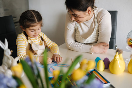 Mom helping her daughter paint an Easter bunny mask. Adorable little girl sitting at the table in the kitchenの写真素材