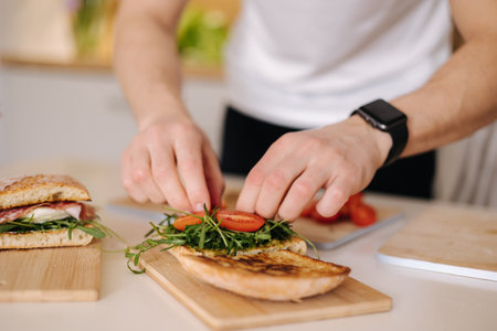 Young man put tomatoes on sandwich. Man preparing lunch at homeの写真素材