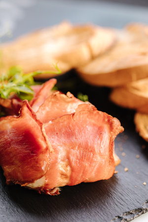 Camembert cheese baked in bacon. Crispy pieces of baguette and slices of chilli on a black stone boardの写真素材