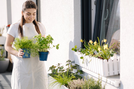 Attractive young female creating garden at balcony. Woman hold pot with dill and basil. Starting for landingの写真素材