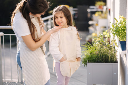 Beautiful young mom helps her daughter to get dressed overall for gardening on balcony. Adorable girl smileの写真素材