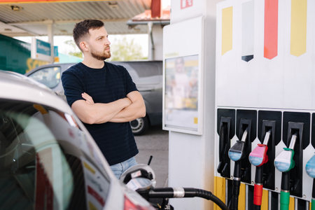 Handsome bearded man cross the arms during refueling car. Male looking on the scoreboard while standing on self service gas stationの写真素材