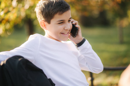Young boy in white sweatshirt with black backpack sitting on the bench in the park and speak with somebody by the phoneの写真素材