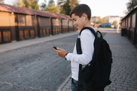 Teenage boy in white sweatshirt and with black backpuck use phone and wireless headphones and print message in social networkの写真素材