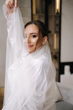 Beautiful bride with long bridal veil dressed in grooms shirt stand in front of big window at homeの写真素材
