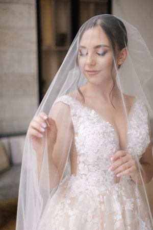 Portrait of gorgeous bride with veil over her face. Wedding day. Woman stand in front of window.の写真素材