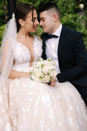 Groom and bride sits on bench and hug each other. Focus on bouquetの写真素材