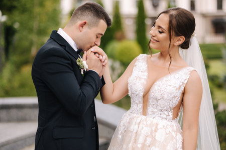 Portrait of handsome groom and gorgeous bride standing outdoor. Bride hold bouquet. Close up of man kiss womens handの写真素材