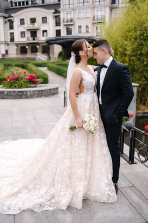 Beautiful bride and groom stand in front of luxury building. Newlyweds go to restaurant to celebrate wedding. Bride kiss groom in forehead.の写真素材