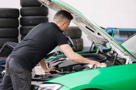 Portrait of a male mechanic working in his garage. Young bearded man checks the engine in the car, the hood is openの写真素材