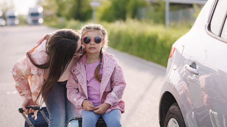 Close-up portrait of mom kiss her daughter in cheek during they waiting a trip. Little girl sitting on her suitcaseの写真素材