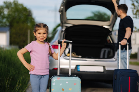 Dad and daughter are standing with their suitcases near a car with an open trunk. Happy little girl preparing to travel.の写真素材
