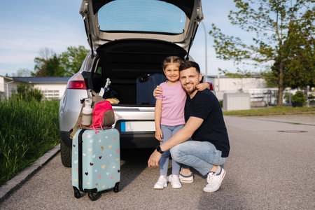 Dad and his daughter stand by the open trunk of their car and preparing for journey while mom turns on the alarm at homeの写真素材