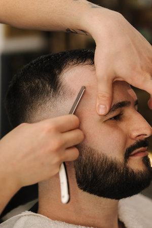 Side view of barber using sharp blade for shaving. Handsome customer sitting in barbershop
