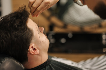 Process of threading procedure in barber shop. Professional barber correcting shape of brows with threads male client sitting in chair.の写真素材