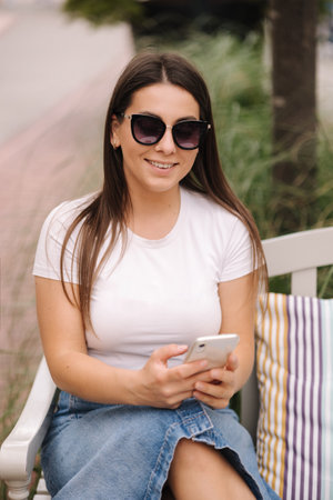 Portrait of beautiful woman sitting on the bench outdoor and using phone. Female in sunglassesの写真素材