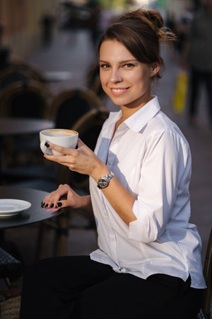 Happy smiled woman drinking coffee on terrace in cafe. Female in white shirt have rest during the breakの写真素材