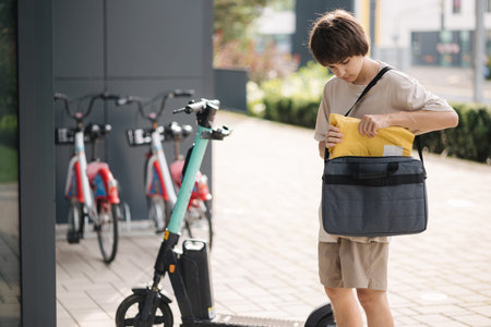 Delivery boy stand by electric scooter and takes the package out of his bag.