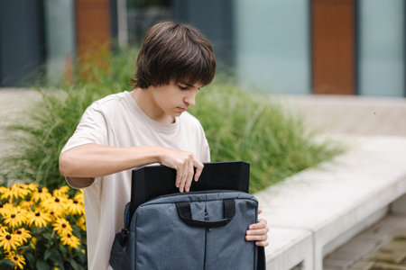 Boy open laptop bag and takes out his laptop. Boy sits on bench outdoorsの写真素材