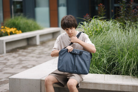Teenage boy sits on bench and open his laptop bag