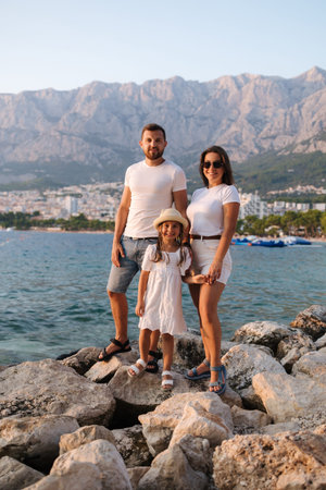 Happy family of dad mom and daughter stand on rocks and looks at the sea. Back view of big mountains