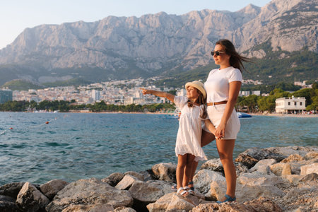 Beautiful mom and daughter stands on rocks and look at the sea. Back view of big mountainsの写真素材