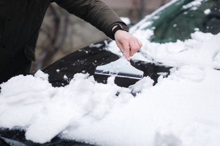 Close up of man is cleaning snowy window on a car with snow scraper. Focus on the scraper. Cold snowy and frosty morning. Black carの写真素材