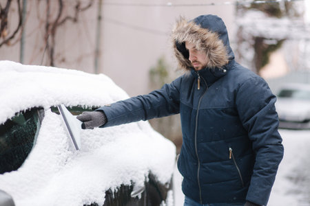 Man in winter jacket clean his car from snow. Winter outdoors. Preparing for drivingの写真素材