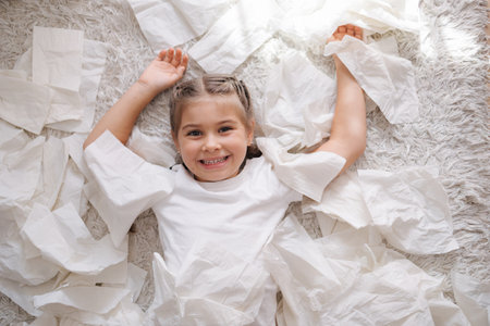 Top view of little girl lies on the carpet among scattered napkins. Girl look into the cameraの写真素材