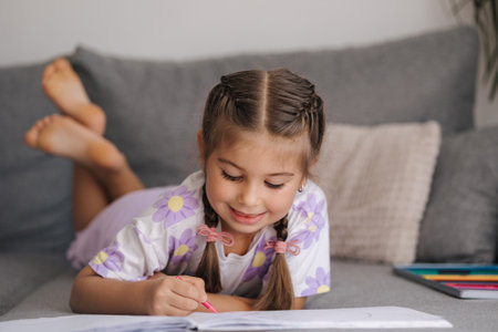 Adorable little girl with beautiful braid hairstyle lying on sofa at home and drawing picture for momの写真素材