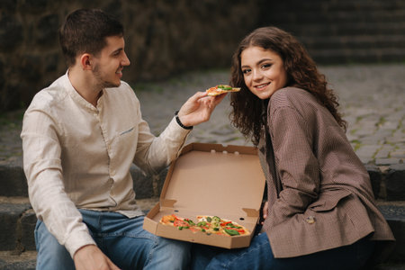 Smiling love couple eating vegan pizza at street on stairs. Handsome man give slice of pizza to womanの写真素材